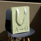 Front view, luxury green Paper Bag Mockup with a white handle and black typographic design on it standing on the navy chair with wooden legs against beige wall.