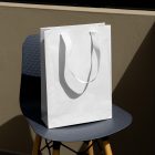 Front view, luxury White Paper Bag Mockup, standing on the navy chair with wooden legs against beige wall.