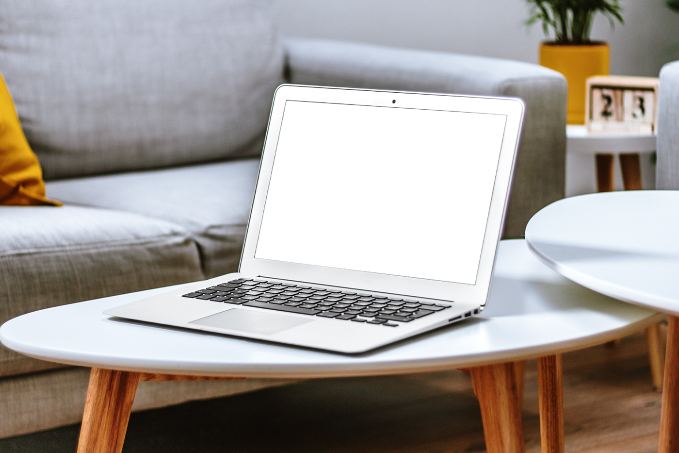 Front view, MacBook Pro Mockup with white screen laying on the white wooden table in front of the sofa.