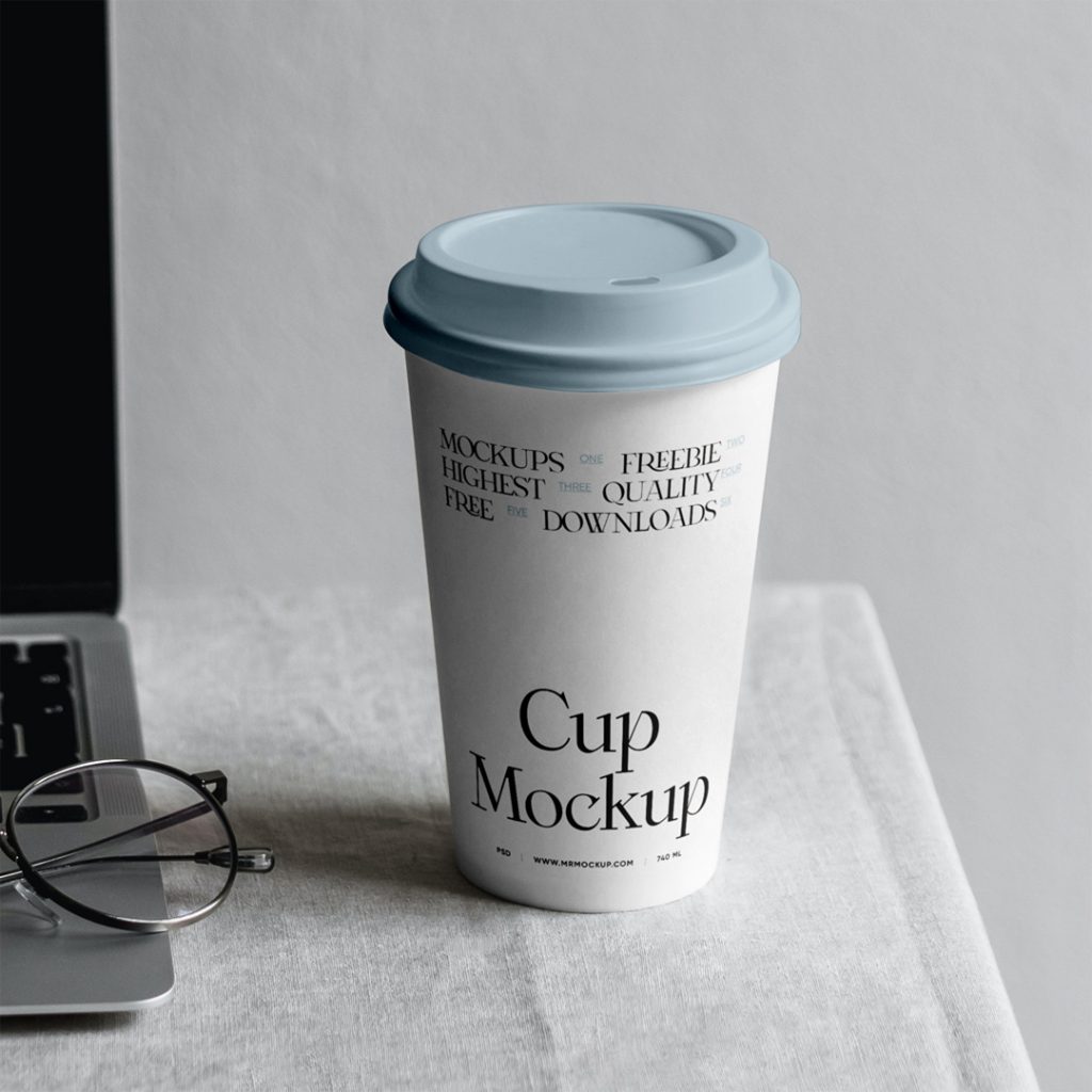 Front view, paper coffee cup mockup standing on the white table right next to the MacBook Pro.