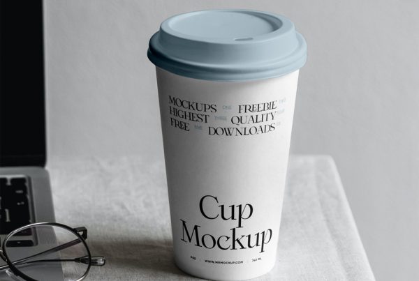 Front view, paper coffee cup mockup standing on the white table right next to the MacBook Pro.