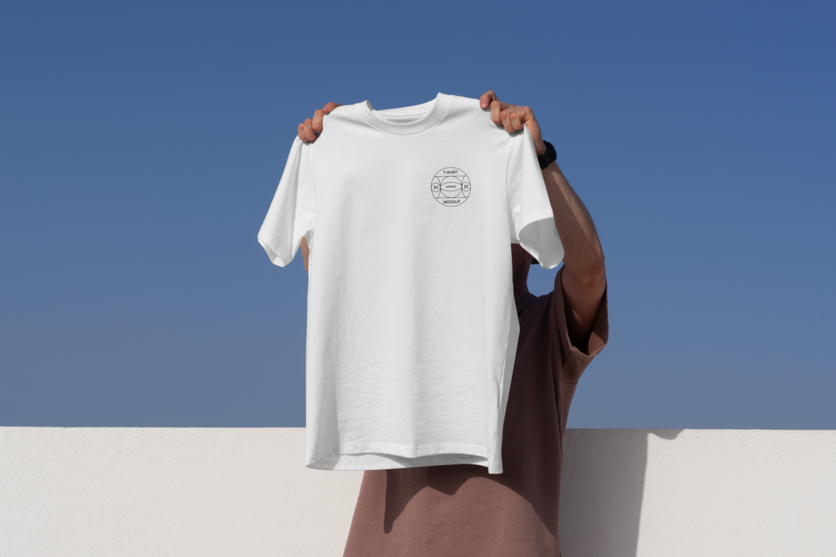 Man holding white t-shirt with small typographic design in front of blue sky.