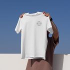 Man holding white t-shirt with small typographic design in front of blue sky.