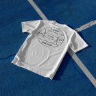 White t-shirt with big black typographic design laying on blue court.