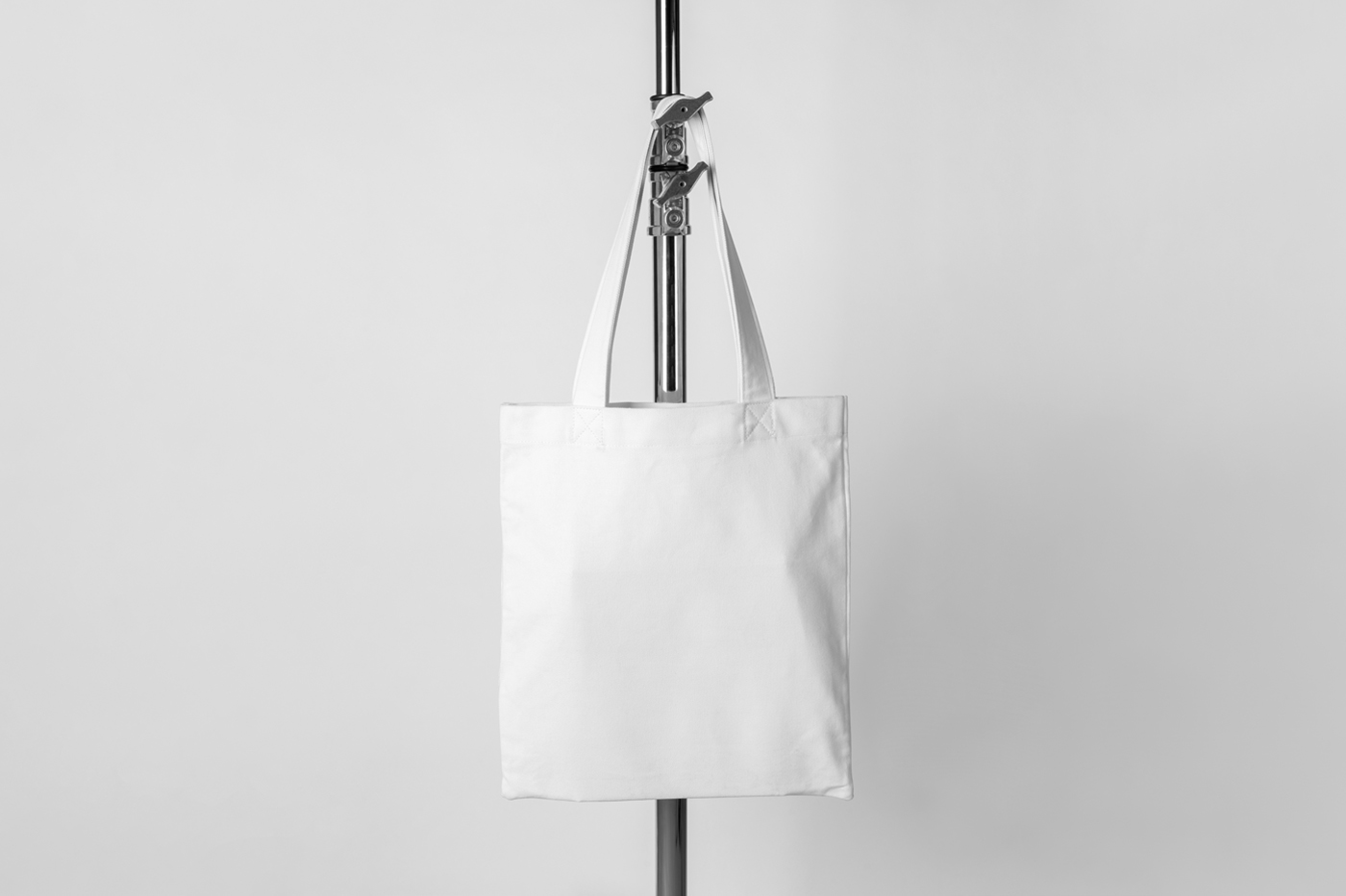 White canvas bag mockup hanging on a tripod in front of the white wall.