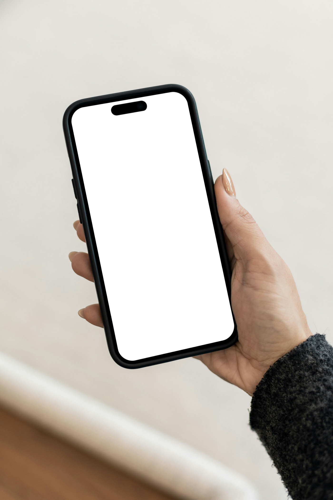 Woman holding iPhone Pro Mockup with white screen in hand in front of the bright beige wall.