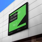 Horizontal city billboard mockup with black, green design and black coffer attached to the bright gray building facade.