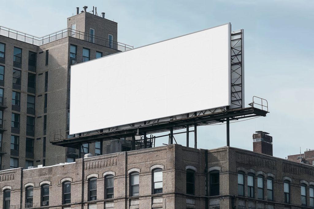 A white city billboard mockup attached to the roof of the building, with the clean blue sky in the background.