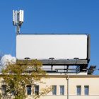 Front view, white city billboard mockup attached to the roof of the building, with the clean blue sky in the background.