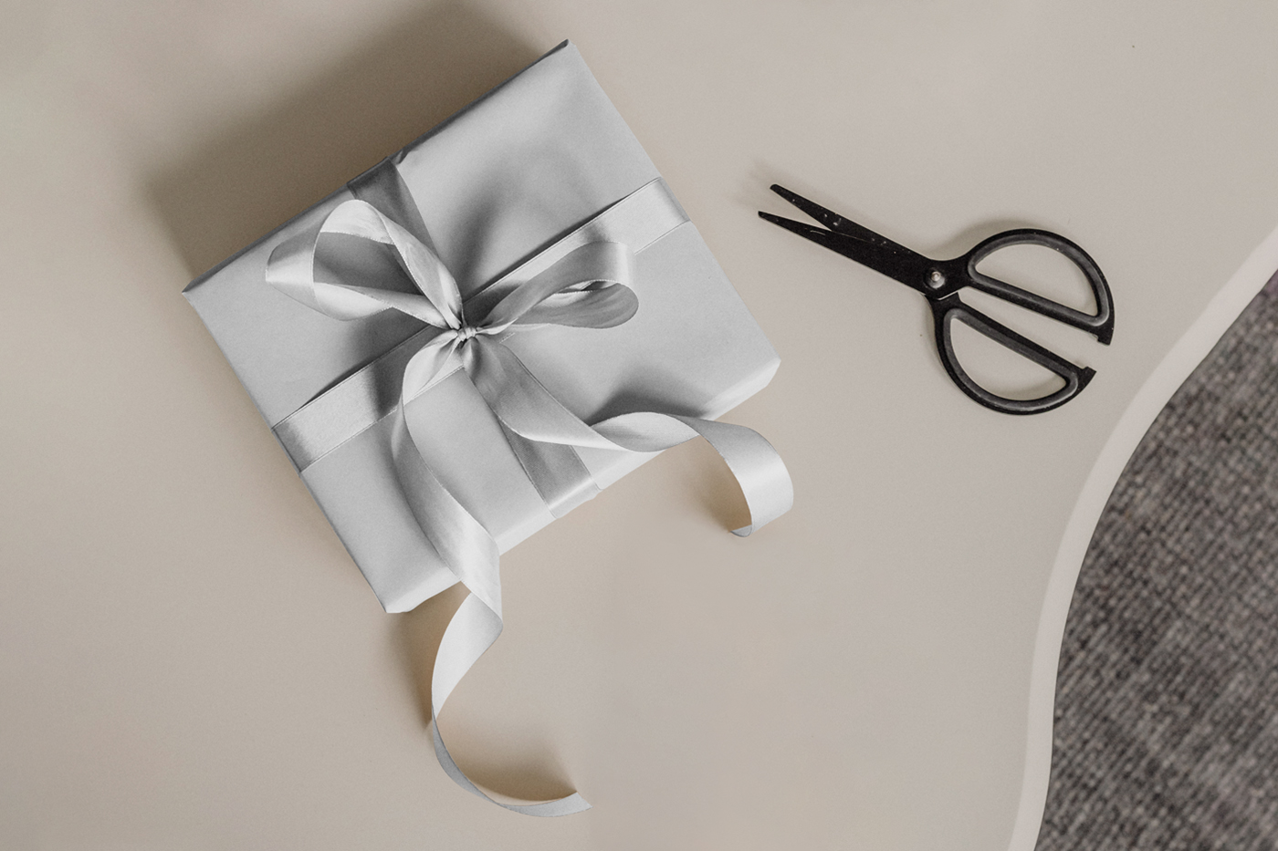 View from above, white gift box mockup with white ribbon laying on the beige table.