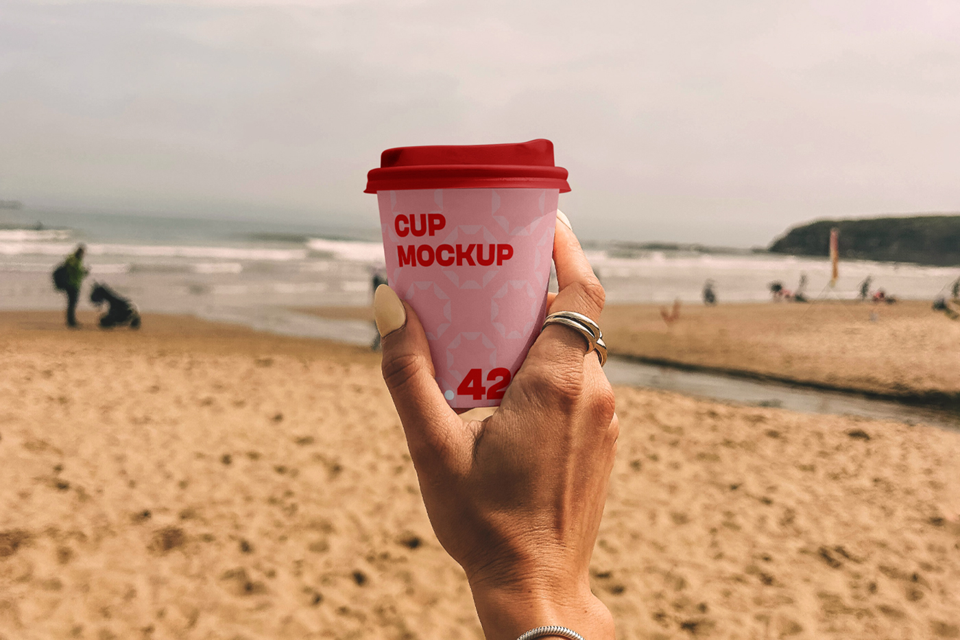 A woman holding a pink paper coffee cup mockup against the background of a sunny beach.