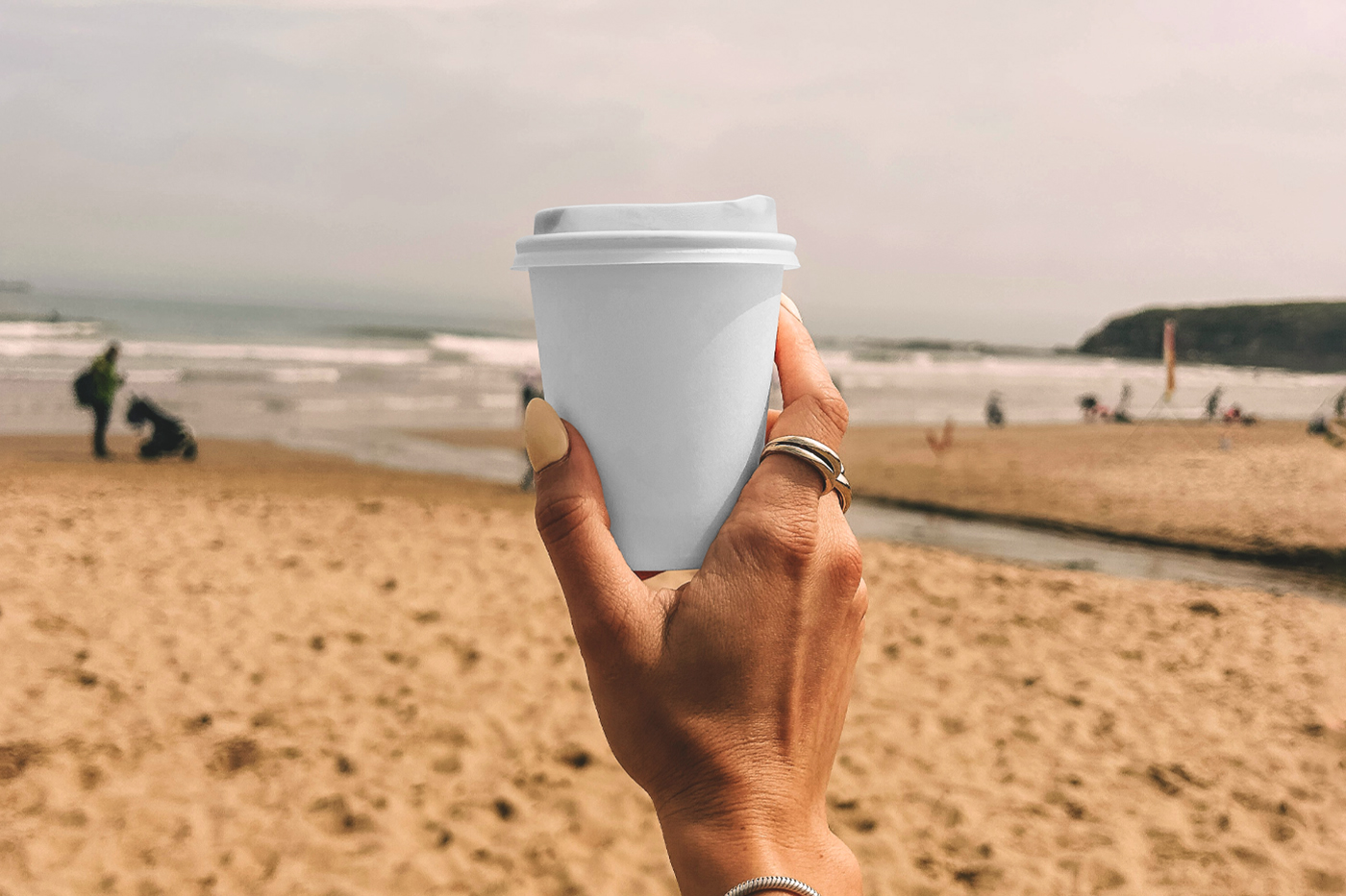 A woman holding a white paper coffee cup mockup against the background of a sunny beach.