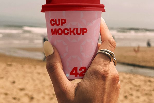 A woman holding a pink paper coffee cup mockup against the background of a sunny beach.