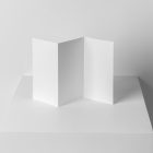 Front view, from above, with a little angle of a single white Trifold Mockup arranged in the letter z standing on a white box. Behind the white box is a huge white wall.