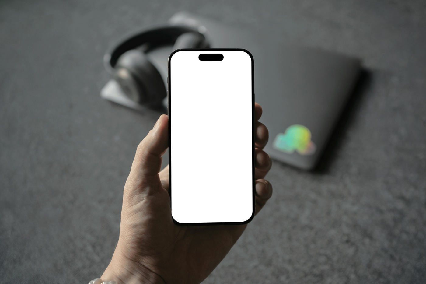 Front view, Hand Holding iPhone Mockup with white screen and visible blurred headphones and laptop laying on the dark gray carpet behind.
