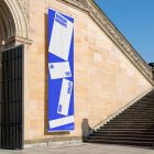 Front view, Vertical Flag Mockup with white and blue design attached to the old architecture building wall. Scene from our Museum Mockups product.