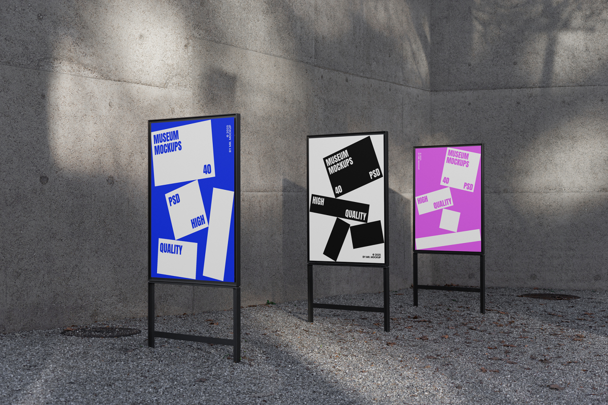 Front view, Three Poster Mockups with blue, white, pink, and white designs and metal frames in black color. Standing in the corner before the concrete wall. Scene from our Museum Mockups product.