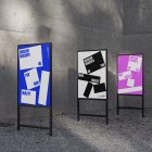 Front view, Three Poster Mockups with blue, white, pink, and white designs and metal frames in black color. Standing in the corner before the concrete wall. Scene from our Museum Mockups product.