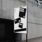 Front view, Poster Mockup with white and black design holding on a black metal pole. Standing against the background of a gray concrete wall. Scene from our Museum Mockups product.