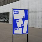 Front view, Poster Mockup with white and blue design holding on a black metal pole. Standing against the background of a gray concrete wall. Scene from our Museum Mockups product.