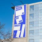 Front view, Vertical Billboard Mockup with white and blue design attached to the modern building against the sky. Scene from our Museum Mockups product.