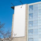 Front view, Vertical White Billboard Mockup attached to the modern building against the sky. Scene from our Museum Mockups product.