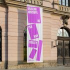 Front view, Hanging Vertical Banner Mockup with white and pink design attached to the silver pole in front of the old building elevation. Scene from our Museum Mockups product.