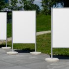 Front view, Three White Poster Mockups standing on the side wall in front of the city park. Scene from our Museum Mockups product.