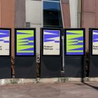 Front view, Five Poster Mockups with purple, black, beige, and neon green geometric designs standing in front of the modern architecture building. Scene from our Museum Mockups product.