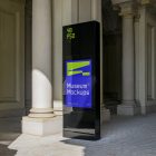 Front view, Monument Poster Mockup with neon green and purple design attached to the sidewalk between old architecture buildings. Scene from our Museum Mockups product.