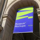View from below, Hanging Banner Mockup with white, purple, and neon green geometric design holding on to ropes attached to the building. Scene from our Museum Mockups product.