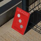 Front view, of outdoor magazine mockups with simple minimalist design in white and red colors, serif typography, and circle foil pattern resists on a metal net fence in black color.