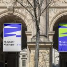 Front view, Two Hanging Banner Mockup with white, purple, and neon green geometric designs holding on to ropes attached to the building. Scene from our Museum Mockups product.