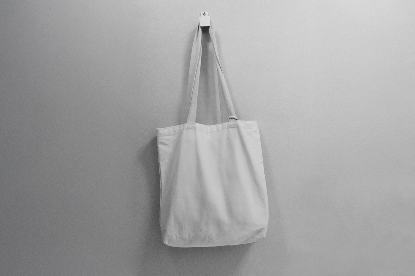 Front view, a white tote bag mockup hanging on a hook attached to the gray color wall.