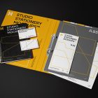 Front view, stationery mockups. Open yellow binder mockup with an A4 card mockup and two business card mockups lying on it, strong geometric typography design in yellow, black, and white colors. Everything is lying on a black background.