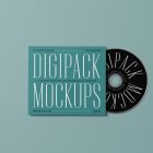 Viewed from above, CD Digipack Mockups presents green disc packaging and a black ejected disc with nice white serif typography lying on the green background.