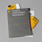 Front view, stationery mockups. A dark grey folder mockup with a protruding yellow leaflet mockup, a white A4 mockup, and a gray square card mockup. Everything is lying on a bright grey background.
