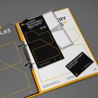Front view, stationery mockups. Open yellow binder mockup with a gray A4 card mockup, black business card mockups, black leaflet mockup, and gray square card mockup lying on it, strong geometric typography design in yellow, black, and white colors. Everything is lying on a dark gray background.