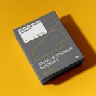 View from above, grey box mockup with a visible white business card mockup lying on it, strong geometric typography design in yellow, white, and black colors. Everything is lying on a yellow background.