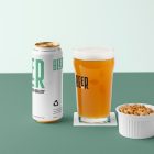 Front view of a standing white beer can mockup with a black and green typography design, a transparent beer glass mockup with a black logo, and a white peanut container standing on the green background floor.