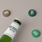 View from above, beer bottle label mockup in white color and black and green typography design with three beer crown caps mockup lying on the beige background.