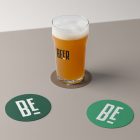 Front view, standing on a brown beer pad, transparent beer glass mockup with black logo design, two green circle beer pad mockups with big white logo design lying on the beige floor.