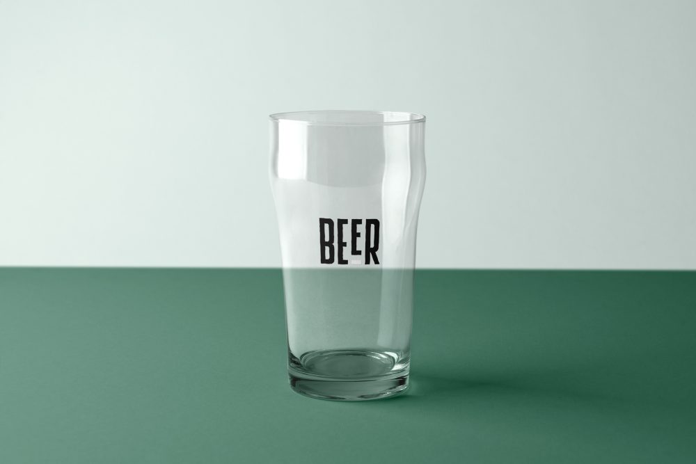 Front view, transparent beer glass mockups with black typography logo design standing on the green floor.