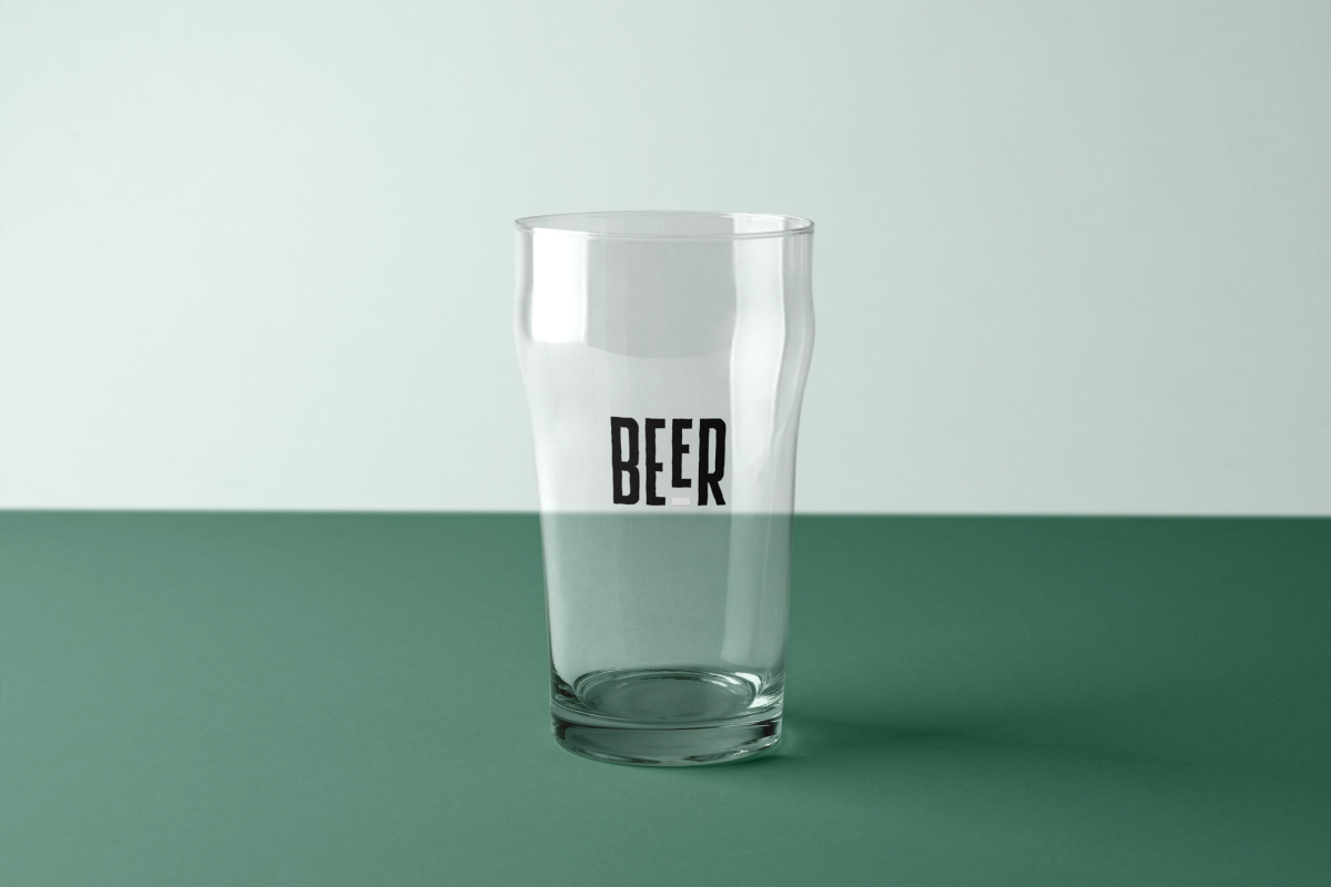 Front view, transparent beer glass mockups with black typography logo design standing on the green floor.