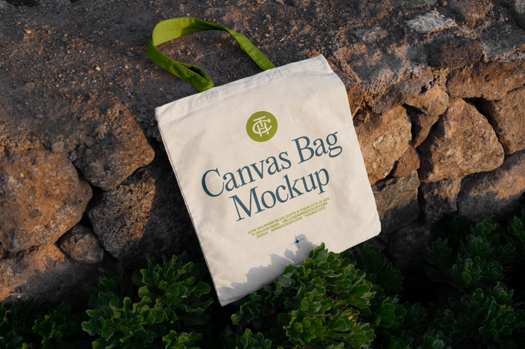 Front view, canvas bag mockup in crème color with a green handle lying on the rocks.
