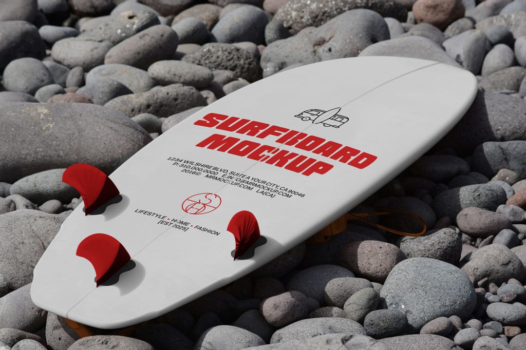 View from above, a white surfboard mockup with a strong red typography design and some red color accents lying on a stone beach.