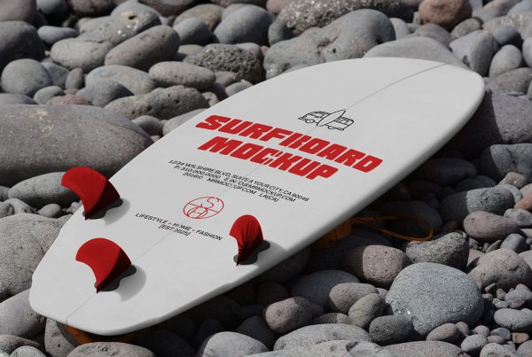 View from above, a white surfboard mockup with a strong red typography design and some red color accents lying on a stone beach.