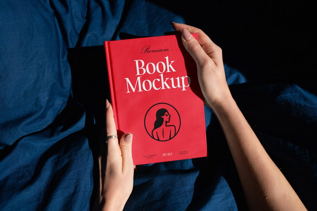 A red book cover mockup in a woman's hands with a white and black design in the background you can see navy-blue bedding.