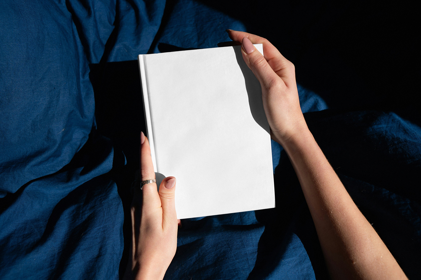 A white book cover mockup in a woman's hands in the background you can see navy-blue bedding.