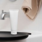 Front view, a white Cosmetic Tube Mockup standing on the black tray. In the background you can see a brightly lit bathroom.